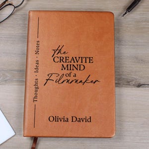 Puede incluir: Un diario de cuero marrón con el texto "The Creative Mind of a Filmmaker" y el nombre "Olivia David" impresos en la portada. El diario también tiene las palabras "Thoughts Ideas Notes" impresas verticalmente.