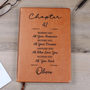 May include: A tan leather journal featuring the text "Chapter 41" and motivational phrases such as "All Your Memories" and "All Your Dreams." The name "Olivia" is also inscribed. The journal is on a wooden surface.