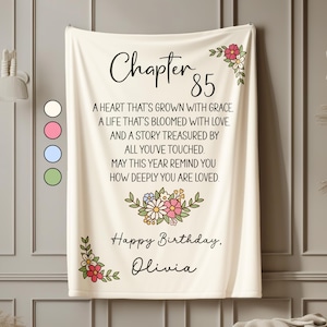 May include: Cream-colored blanket with the text "Chapter 85" and a floral design. The blanket has a personalized message, "Happy Birthday, Olivia." The design includes a variety of flowers in pink, white, and blue.