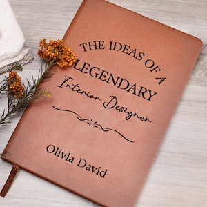 May include: A brown leather-bound journal with the text "THE IDEAS OF A LEGENDARY Interior Designer Olivia David" in black lettering. A decorative sprig of orange flowers rests on the journal's corner. A brown ribbon bookmark is visible.