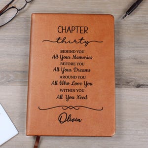 Puede incluir: Un diario de cuero marrón con las palabras "CHAPTER thirty" y texto inspirador. El diario incluye el nombre "Olivia" en la parte inferior. Un bolígrafo y gafas son visibles en el fondo.