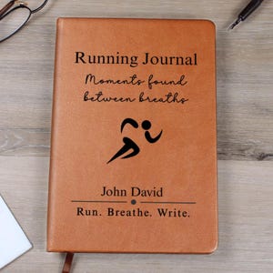 Może przedstawiać: Brązowy, skórzany dziennik biegacza z napisem "Running Journal" i "Moments found between breaths". Pod tekstem znajduje się czarna grafika biegacza. Na okładce znajduje się również imię "John David" i fraza "Run. Breathe. Write."