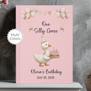 May include: Pink hardcover book with the text "One Silly Goose" and "Olivia's Birthday July 20, 2025". The cover features a watercolor illustration of a goose wearing a party hat and holding a birthday cake, with balloons and bunting.