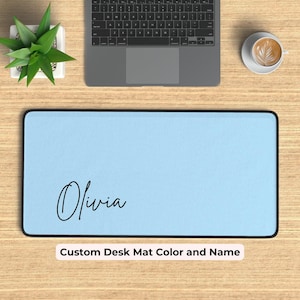 May include: A light blue desk mat with a black border and the name "Olivia" written in black cursive script. The mat is on a wooden surface with a laptop, a plant, and a cup of coffee.