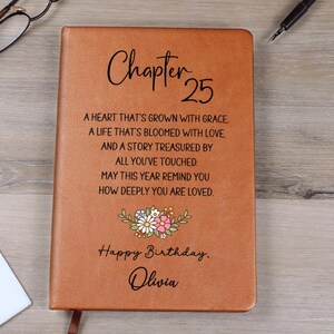 May include: A brown leather-bound journal with the words "Chapter 25" in elegant script. The cover features a floral design and the text "Happy Birthday, Olivia," along with a heartfelt message. A pen and glasses are visible.