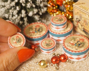 May include: Set of six miniature round boxes with red and white checkered patterns and blue trim. Each box features a different illustration of children in vintage clothing. The boxes are arranged on a white surface with gold and red ornaments.