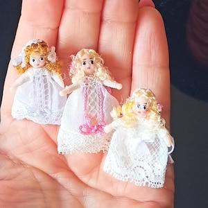 May include: Three miniature dolls with blonde and auburn hair, wearing white dresses with lace details. One doll has a veil. The dolls are held in a hand, showcasing their small size and intricate design. A charming collection.