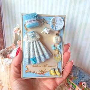 Può includere: Un'esposizione in miniatura a tema baby con un vestito blu e bianco, una coperta blu e un bottone "Hello Peter". Altri oggetti includono un flacone di profumo, un bavaglino e stivali gialli. L'esposizione è su uno sfondo blu con accenti floreali.