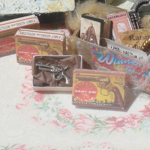 Könnte beinhalten: Miniatur-Silberrevolver in einer kleinen weißen Schachtel mit braunem Papier. Mehrere Schachteln mit "DAISY KID" Spielzeugpistolen sind zu sehen, zusammen mit anderen Ephemera im Vintage-Stil. Die Schachteln sind gelb und rot mit einer Pistolenillustration. Der Hintergrund ist ein Blumenmuster.