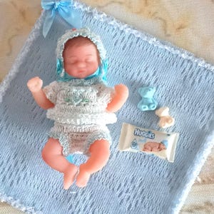 Può includere: Una bambola neonata in miniatura con un completo all'uncinetto bianco e un cappuccio blu, su una coperta all'uncinetto azzurra. Sono presenti anche due piccole figure di orsetti e una confezione di salviettine umidificate Huggies.
