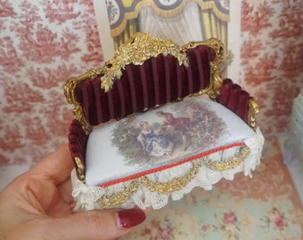May include: A miniature red velvet sofa with gold accents and a floral patterned cushion. The sofa is decorated with lace and a red ribbon.