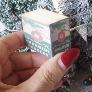 May include: A miniature box of Christmas tree ornament hangers. The box is green and white with red and white ornaments on the front. The text on the box reads "Christmas Tree Ornament Hangers".