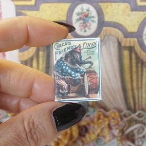 May include: A vintage circus themed card featuring an elephant playing a drum. The card is titled "Circus Friends" with the text "with Pleasure Pictures Series" below. The card is in a hand holding it up.