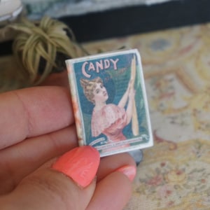 May include: Vintage candy box featuring a woman holding a stick of candy. The box is blue and white with the word "CANDY" printed in large letters. The text on the box reads "Candy, how delicious!" and "Manufactured by William Hill & Sons, Philadelphia, Pa."