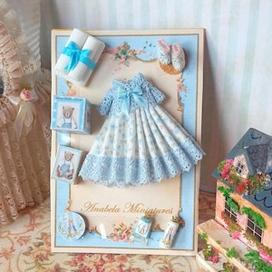 Può includere: Un'esposizione in miniatura con un vestitino, scarpe e accessori per neonati. L'abito è bianco con un motivo floreale blu e rifiniture in pizzo. L'esposizione include anche una scatola di crema per bambini e un cartello con la scritta "Anabela Miniatures."
