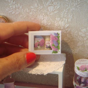 May include: A small white frame with a picture of a lilac flower and a bottle of lotion. The frame is on a white surface with a lace doily.