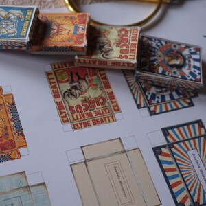Può includere: Scatole a tema circo in miniatura con grafica vintage. Le scatole presentano disegni rossi, blu e gialli, con testi come "CIRCUS MINKENIE" e "CLYDE BEATTY CIRCUS". Sono visibili anche modelli stampati per le scatole.