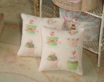 Op de afbeelding: Twee roze en witte decoratieve kussens met een cupcake-ontwerp. De kussens hebben verschillende cupcakes met roze, groene en witte frosting.