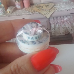 Può includere: Una torta in miniatura a due piani decorata con un nastro rosa e il viso di un bambino. Il piano superiore ha la scritta "PARIS" stampata.