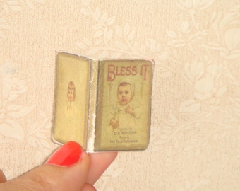 以下が含まれることがあります： 表紙に子供が描かれた「Bless It」というタイトルのヴィンテージブック。白いドレスと白いボンネットを着た子供が描かれています。この本は、茶色の紙に金色の背表紙で製本されています。