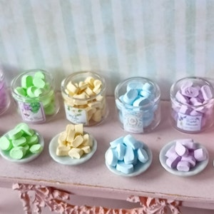 May include: Eight glass jars filled with pastel-colored candy, arranged in a row on a shelf. Each jar has a label with a floral design.  There are also eight small plates with the same colored candy on them.