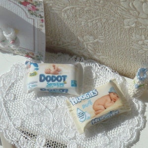 Może przedstawiać: Zbliżenie miniaturowych artykułów dla niemowląt. Widoczne są dwa opakowania pieluch, jedno z napisem "DODOT Sensitive", a drugie "Huggies Pure". Para małych szydełkowych bucików dla niemowląt w kolorze białym i jasnoniebieskim. Na białej koronkowej serwetce.