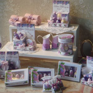 May include: A miniature dollhouse display of lilac blossom-themed bath and body products. The display features a variety of bottles, jars, and boxes, all decorated with lilac blossoms and the words "Lilac Blossom". The display is set up on a white shelf with a lace tablecloth.