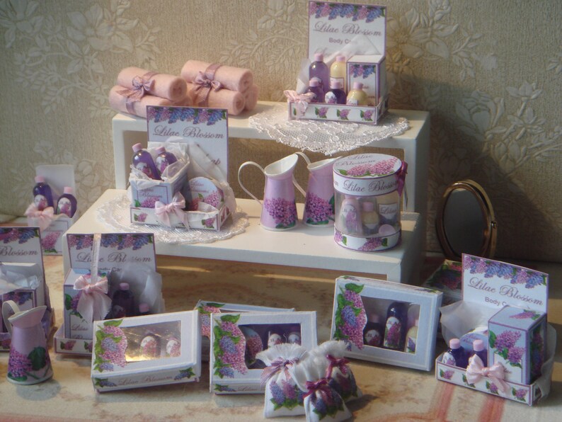 May include: A miniature dollhouse display of lilac blossom-themed bath and body products. The display features a variety of bottles, jars, and boxes, all decorated with lilac blossoms and the words "Lilac Blossom". The display is set up on a white shelf with a lace tablecloth.