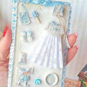 Può includere: Un'esposizione in miniatura a tema baby con un vestito di pizzo bianco, bavaglino, stivaletti e cappellino. L'esposizione include un sonaglio, un flacone piccolo, un ciuccio, un cavallo a dondolo e un piatto. La cornice è rifinita con un nastro blu.