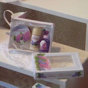 May include: A set of three miniature bath products in a white box with a lilac floral design. The box says "Lilac Blossoms" and contains a bottle of body wash, a bottle of lotion, and a small bottle of bath salts.