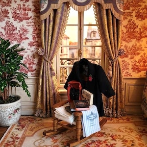 Può includere: Una sedia a dondolo in miniatura in legno sostiene un cappotto nero con una spilla floreale rossa, due piccoli libri, un panno bianco piegato e un asciugamano bianco con testo blu. La scena è ambientata in una stanza con carta da parati toile.