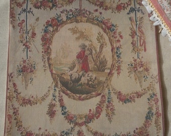May include: A beige tapestry with a central oval design featuring a shepherdess with sheep in a landscape setting. The oval is surrounded by garlands of pink, blue, and green flowers.
