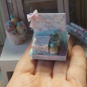 May include: A miniature gift set with a pink and white floral design. The set includes three small bottles of perfume, a box of soap, and a lavender sachet. The set is displayed on a hand.