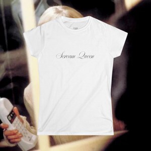 May include: White t-shirt with the text "Scream Queen" in a cursive font. The shirt has a crew neck and short sleeves. The image is taken in a dimly lit setting, with a person and a phone in the background.