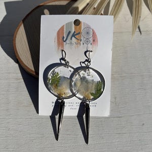 May include: A pair of circular resin earrings featuring embedded dried white flowers and green foliage. Each earring hangs from a silver-toned hook and is accented with a long, pointed, metallic black spike charm dangling from the bottom edge.
