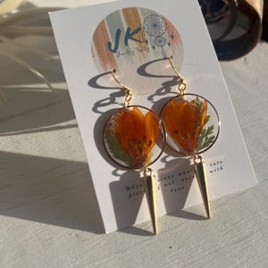 May include: Gold-tone earrings with orange flower petals and green leaves encased in clear resin. Each earring has a gold spike pendant. The earrings are displayed on a white card with the text "JK Jewelry Kelly".