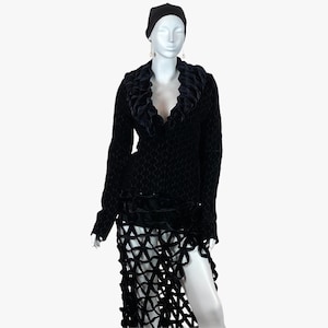 May include: A black ensemble on a mannequin. The outfit includes a textured, long-sleeved top with a ruffled collar and a skirt with a geometric cutout design. A black beanie and earrings complete the look. The fabric appears to be velvet.