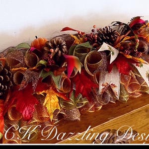 May include: A fall-themed table runner with a mix of orange, red, brown, and gold mesh, pinecones, faux leaves, and pumpkins. The runner is on a wooden surface with the text "CK Dazzing Design" visible in the bottom right corner.