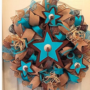 May include: A turquoise and brown star-shaped wreath with burlap and brown ribbon accents. The stars have a brown outline and a silver concho in the center.