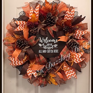 May include: A brown, orange, and white fall wreath with a wooden sign that reads "Welcome and be blessed all who enter here". The wreath is decorated with pine cones, fall leaves, and orange and white chevron ribbon.