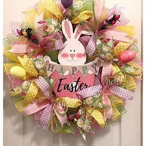 May include: A pink and yellow Easter wreath with a bunny and a sign that says "Happy Easter". The wreath is decorated with ribbons, mesh, and eggs.
