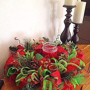 May include: A red and green Christmas centerpiece with a red candle in a glass jar. The centerpiece is made of mesh ribbon, pine sprigs, and pine cones. The centerpiece is on a wooden table with two candle holders in the background.
