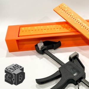 May include: Orange plastic ruler with markings in millimeters, a black clamp, and a 3D printed orange box with the text "EMB 3D" on the side.