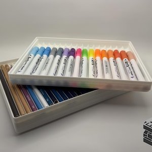 May include: A white tray filled with colorful markers, including blue, gray, purple, pink, green, yellow, orange, and red. Another tray below holds pencils. The markers are labeled "Arrtx Sketch Markers". The image has a white background.