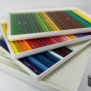 May include: A collection of colored pencils arranged in white trays. The pencils are organized by color, ranging from blues and greens to reds and yellows. The trays are stacked, showcasing the variety of colors available.