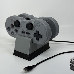 May include: A gray video game controller charging station with two controllers. The controllers have black buttons and a cross-shaped directional pad. A black USB-C cable is connected to the base. The logo "EMB 3D" is in the bottom right corner.