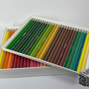 May include: Two white rectangular trays filled with colored pencils. The pencils are arranged in rows by color, ranging from yellow to red, green, and brown. The pencils are neatly organized, creating a visually appealing display.