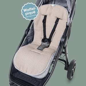 Può includere: Un rivestimento per sedile di passeggino beige a trama waffle con un'imbracatura nera è mostrato all'interno di un passeggino nero. Il rivestimento ha un'etichetta che recita "Waffel-piqué 100% Baumwolle". Il passeggino ha quattro ruote.