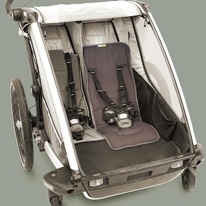 Könnte beinhalten: Ein grauer und weißer Doppelkinderwagen mit zwei Sitzen, jeweils mit Sicherheitsgurt. Der Kinderwagen hat eine Seitenwand aus Mesh, einen Lenker und zwei Räder. Konzipiert für den Außeneinsatz.