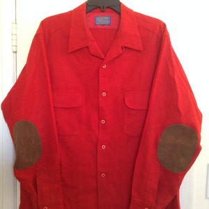 Könnte beinhalten: Ein rotes Langarmhemd mit braunen Leder-Ellenbogenpatches und einer Knopfleiste vorne. Das Hemd hat zwei Taschen vorne und ein blaues Etikett am Kragen mit der Aufschrift "Pendleton Woolen Mills".